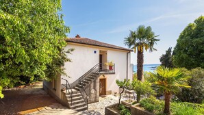 Un bâtiment à deux étages, doté d’un escalier menant à un balcon, entouré d’une végétation luxuriante et offrant une vue dégagée sur la mer.