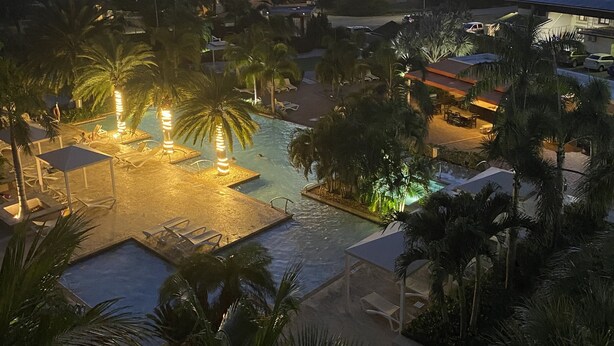 Ein Resort mit Swimmingpool, umgeben von Palmen und Liegestühlen, mit Blick auf ein Gewässer und Gebäude im Hintergrund.