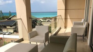 Ein Balkon mit einer beigen Couch, Sesseln und einem Glasgeländer mit Blick auf Strand und Meer.