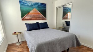 A bedroom with a bed, a ceiling fan, a wooden floor, a painting of a pier, and a mirror. - Courtyard Villa with gas golf cart located between two Town Squares! (The Villages)