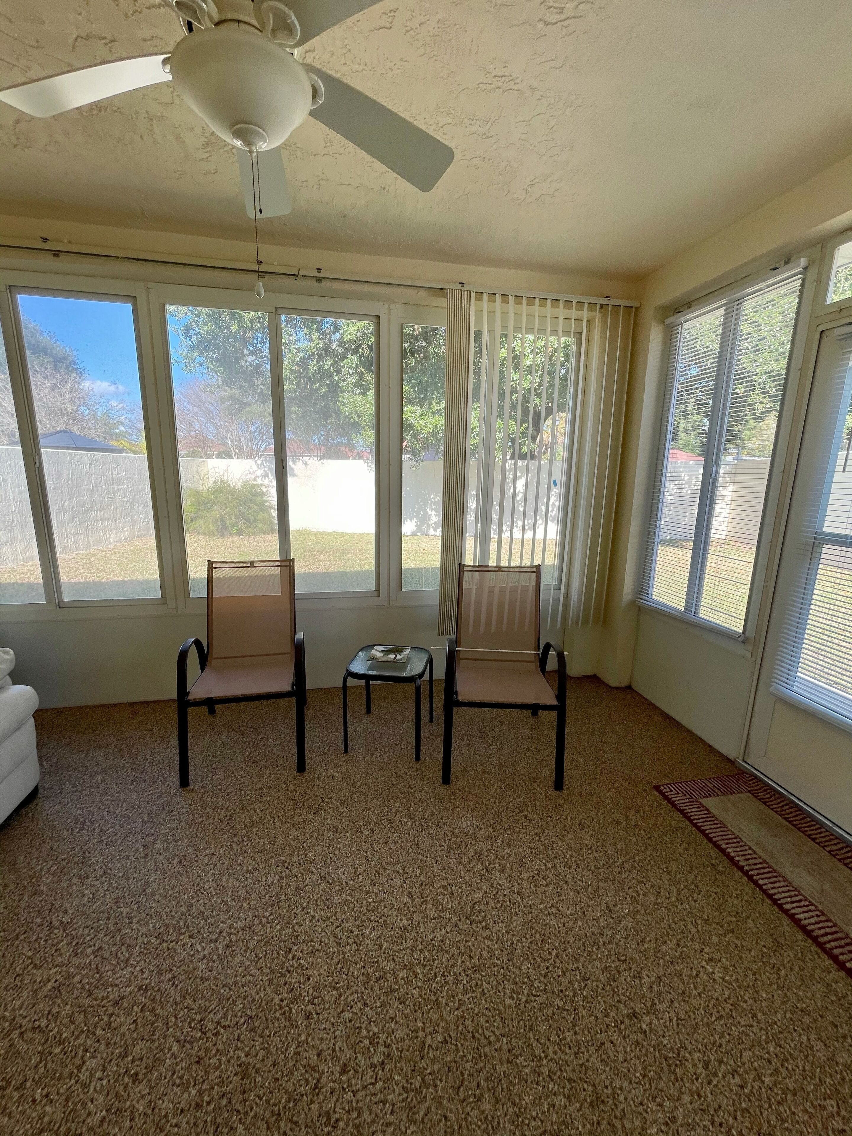 A room with a ceiling fan, large windows, a carpeted floor, and two chairs.