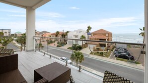 A balcony with wicker furniture overlooking a street with parked cars and houses, with a view of the ocean.