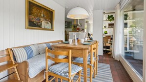 Uma área de jantar acolhedora com uma mesa de madeira, cadeiras e um banco. Um quadro está pendurado na parede, e uma janela grande oferece luz natural.