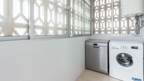 A laundry room with a compact washer and dryer, large windows with patterned blinds, and a visible water hose. - Gorgeous apartment in Santa Pola with WiFi (Santa Pola)