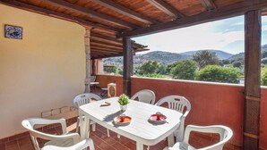 A covered outdoor patio with a table and chairs, a potted plant, and a view of a mountainous landscape.