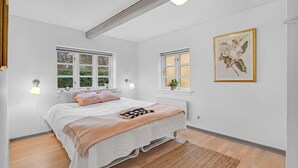 Un dormitorio con una cama, dos ventanas, una lámpara de pared, una impresión botánica enmarco y suelo de madera.