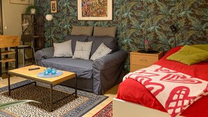 A room with a grey sofa, striped pillows, a small table with a remote and glasses, a red bed with a patterned blanket, and a wall with a leafy wallpaper. - Lovely apartment in Borgholm with WiFi (Borgholm)