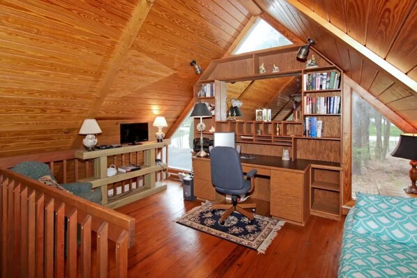 Une chambre confortable située sous les toits, avec un plafond en pente en bois, un bureau avec une chaise, une bibliothèque et un téléviseur.