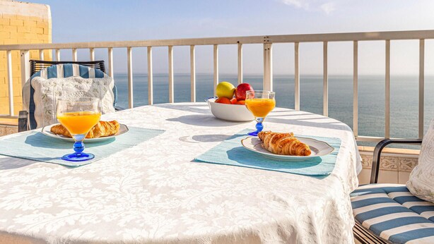 En balkon med et bord dækket til morgenmad, med croissanter, frugt og appelsinjuice, med udsigt over havet.