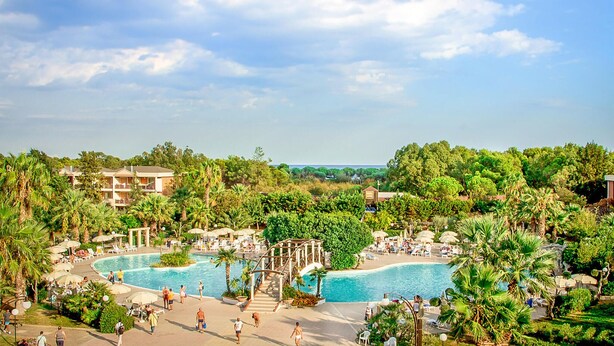 A resort with two large swimming pools surrounded by palm trees and lounge chairs. There are multiple buildings with balconies and a clear view of the ocean.