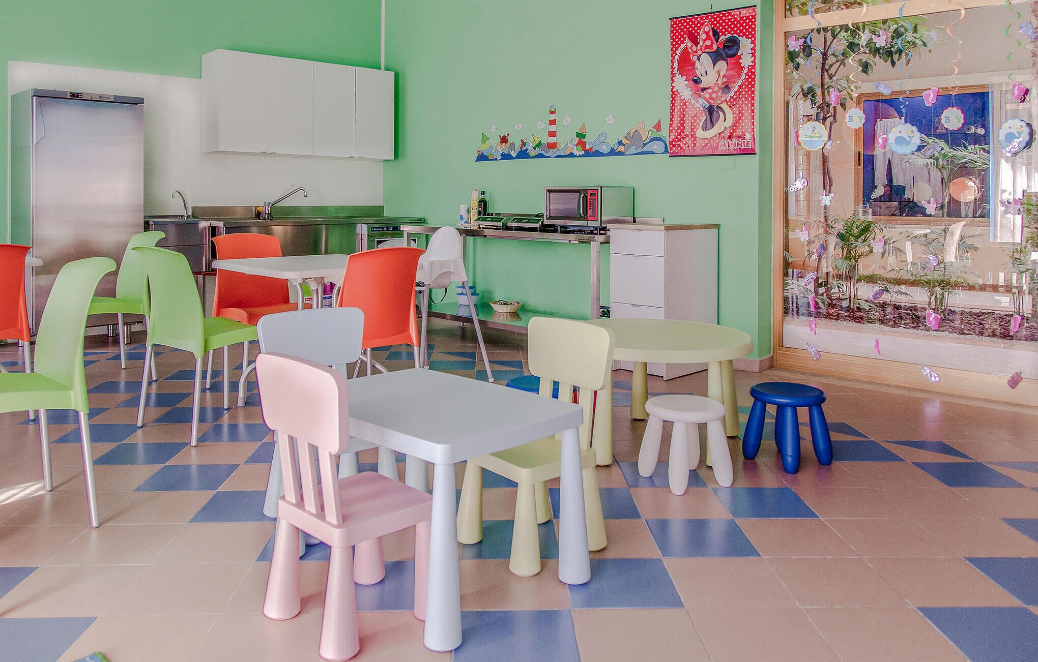 Una sala de jocs infantil amb cadires i taules de colors, un frigorífic, un microones i una finestra amb decoracions.