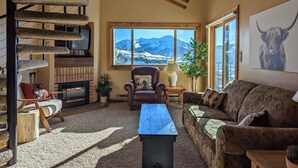 A cozy living room with a fireplace, a comfortable sofa, a wooden coffee table, and a view of mountains through a large window.