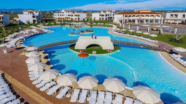 A large swimming pool with multiple sun loungers and umbrellas, surrounded by buildings with a Mediterranean architectural style.
