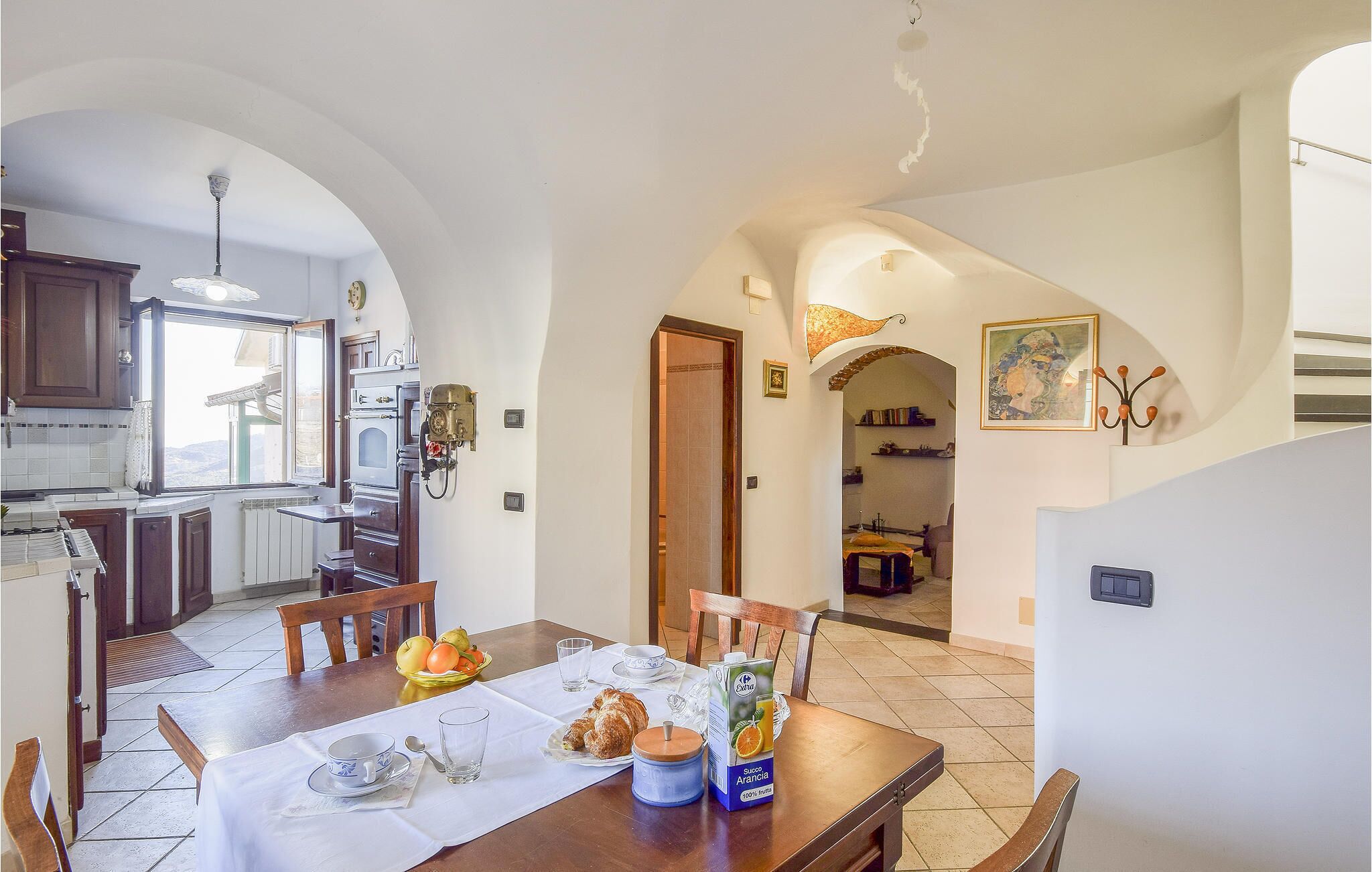 A dining area with a table set for two, a kitchen visible through an archway, and a dining room with a painting on the wall.