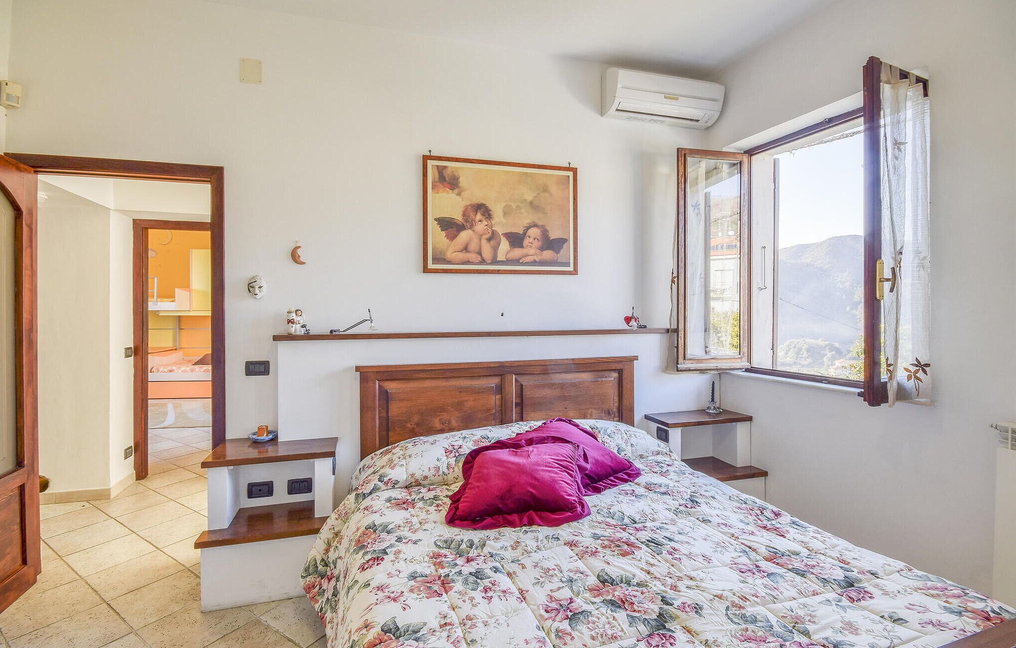 A bedroom with a wooden bed, a painting above the bed, a window with a view, and a small bedside table.