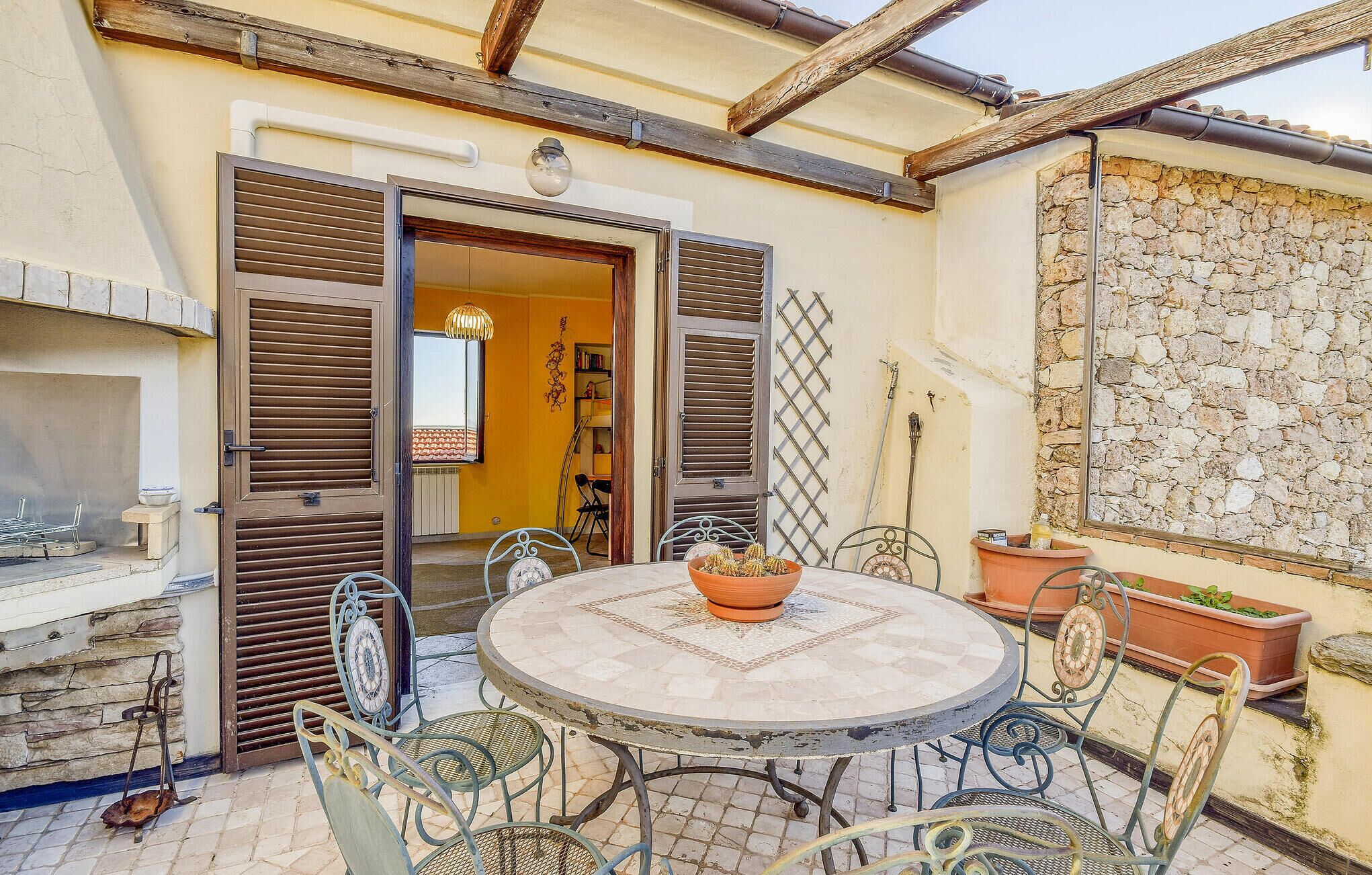An outdoor patio with a round table and metal chairs, a stone wall, and a wooden ceiling.
