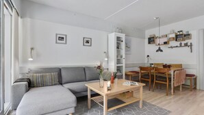 A modern living room with a grey sectional sofa, a wooden coffee table, and a dining area with a wooden table and chairs. There are wall-mounted shelves with decorative items and a white cabinet with glass doors. - Stunning apartment in Hasle (Hasle)