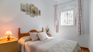 A bedroom with a wooden bed, a nightstand with a lamp, a window with floral curtains, and a wall-mounted artwork. - Pet friendly home in Sayalonga (Sayalonga)