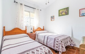 Een slaapkamer met twee houten bedden, een nachtkastje met een lamp, een raam met gordijnen, en twee ingelijste foto’s aan de muur.
