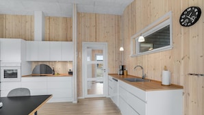 A modern kitchen with wooden walls, white cabinets, a wooden countertop, a sink, a clock, and a dining area with a table and chairs.