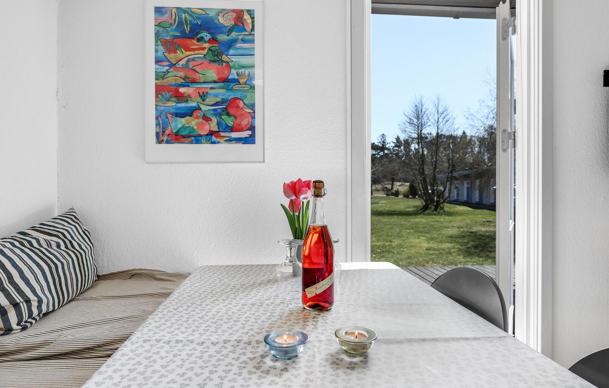 A room with a white table, a colorful painting, a bottle of red liquid, and a view of a grassy area outside.