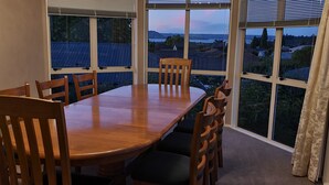 A dining room with a wooden table and chairs, large windows with a view of houses and trees, and a curtain on the right. - Large & Luxurious Family Home in a Private and Peaceful area  (Rotorua)