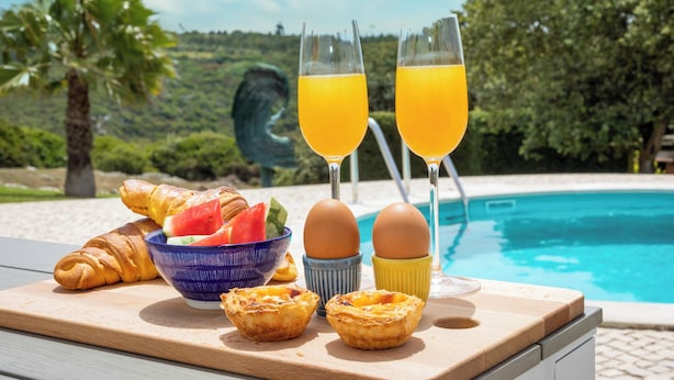 A breakfast setup with croissants, a bowl of fruit, eggs, and two glasses of orange juice by a pool with a scenic view. - Villa with private pool and large garden (Ribamar)