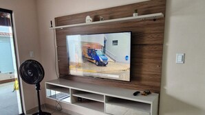 Een woonkamer met een flatscreen televisie aan een houten wand, een witte meubelunit eronder en een ventilator aan de linkerkant.