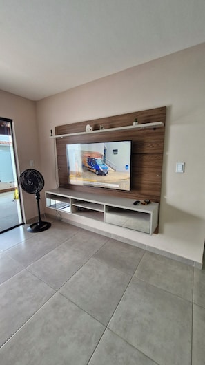 Un salon avec un téléviseur à écran plat fixé à un mur en bois, un meuble de rangement blanc en dessous et un ventilateur sur pied à gauche.
