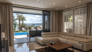 Una sala moderna con un amplio sofá modular, una mesa de centro de madera y vistas a una piscina y zona exterior con plantas y sillas.