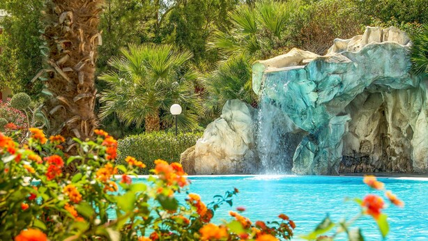 A pool with a waterfall feature surrounded by lush greenery and flowering plants. - Lovely apartment in Nova Siri MT (Nova Siri MT)