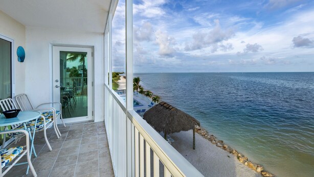Un balcon avec vue sur la mer, une table avec des chaises et un parasol en chaume.