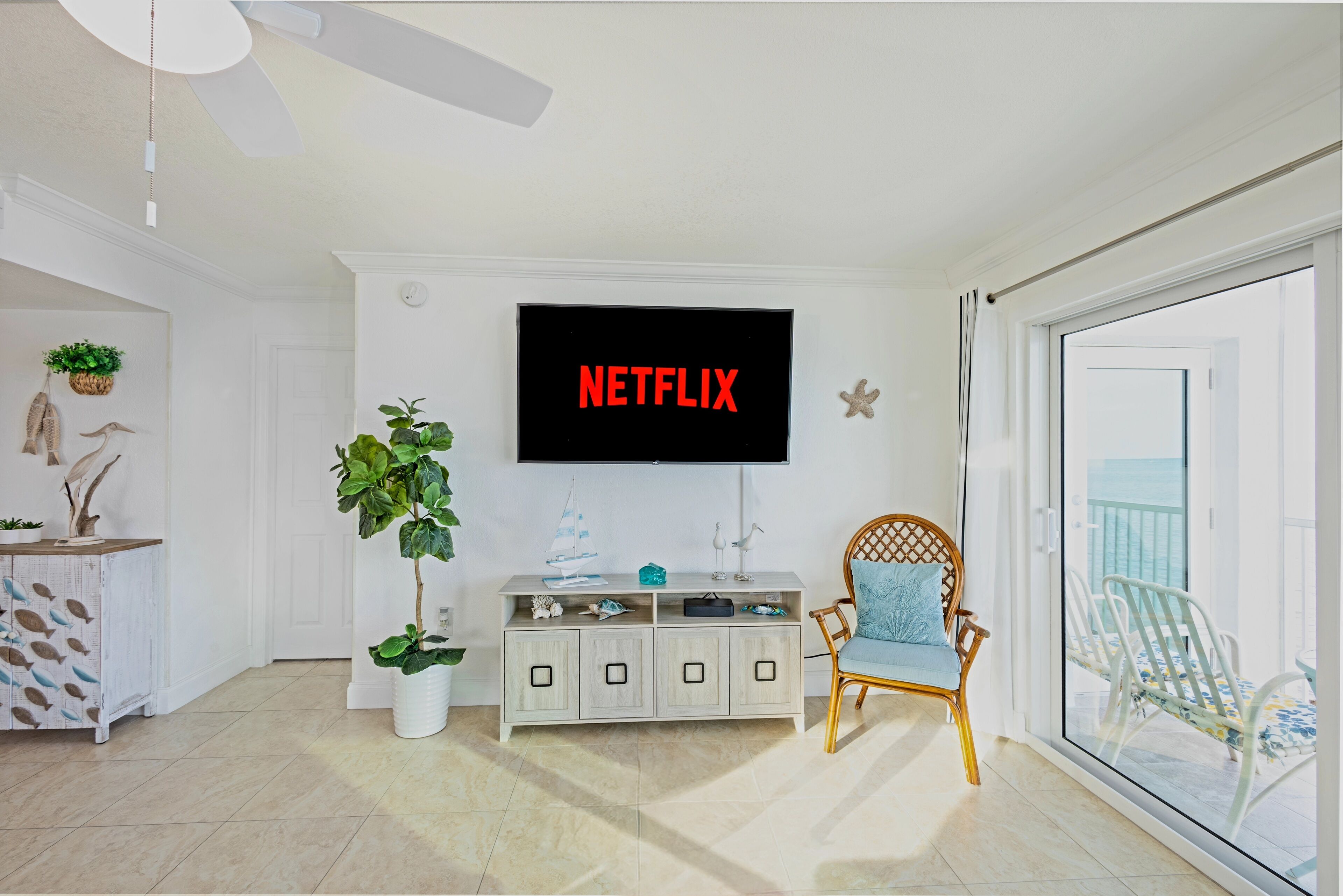 Un salon avec un téléviseur affichant le logo « NETFLIX », un meuble blanc, un fauteuil en osier et une plante en pot.