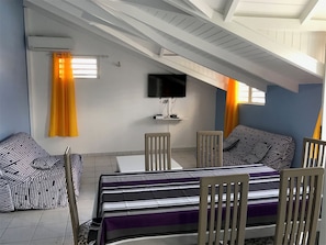 A room with a dining table, chairs, a TV mounted on the wall, and a bed with striped bedding.