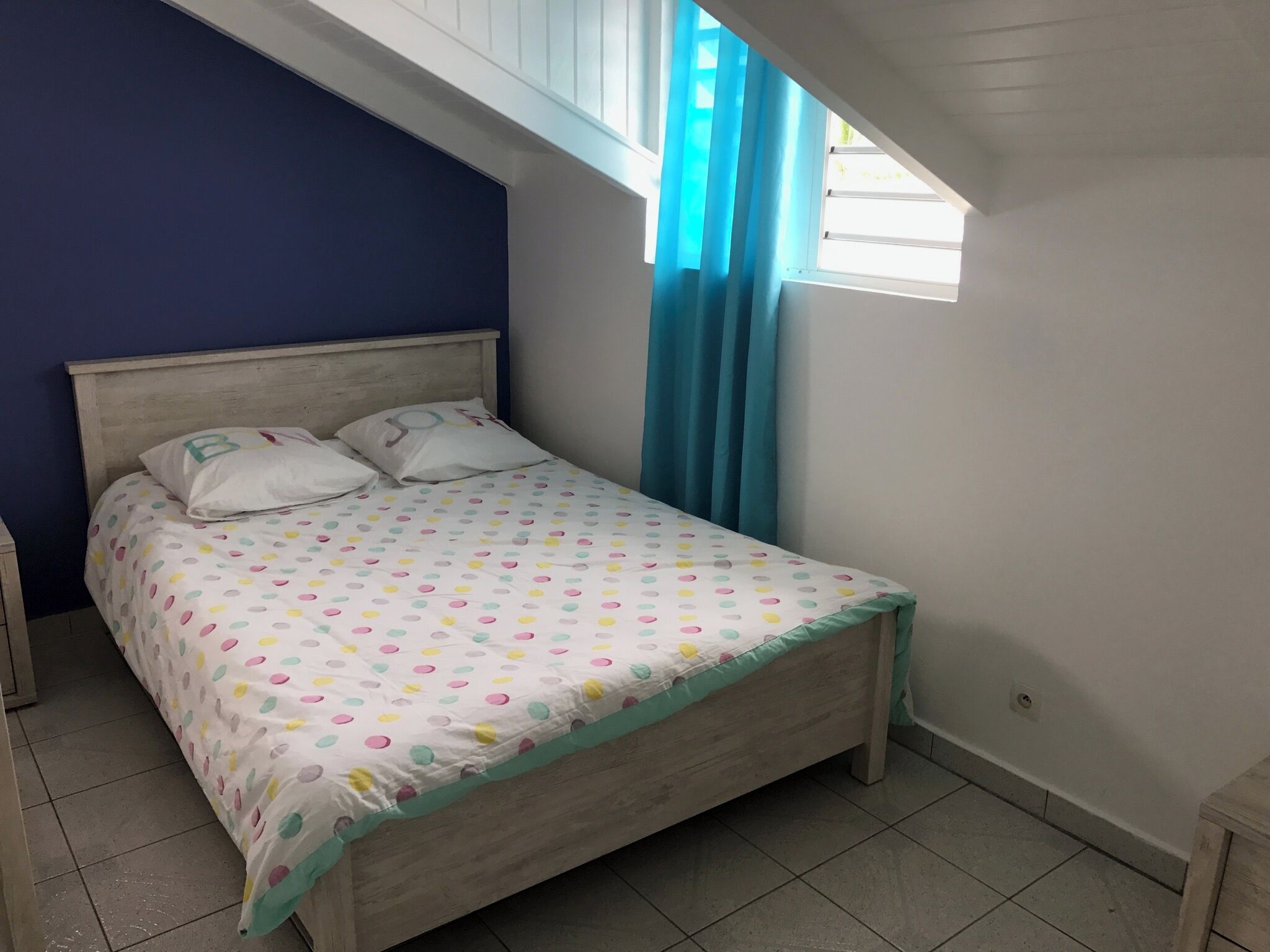 Ein Schlafzimmer mit einem Bett, einem Nachttisch, einem Fenster mit Vorhängen und einer geneigten Decke.
