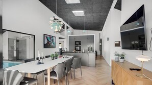 Uma cozinha moderna com uma ilha central, uma área de refeições com mesa e cadeiras brancas, e uma televisão grande fixada na parede.