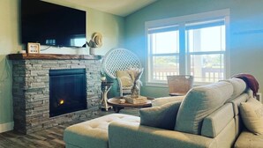 Una sala de estar con chimenea de piedra, un televisor de pantalla plana montado encima y un sofá cómodo, con un amplio ventanal que proporciona luz natural.