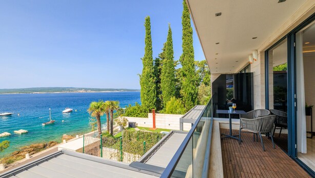 Un balcon avec vue sur la mer, de grands arbres et une table avec des chaises.