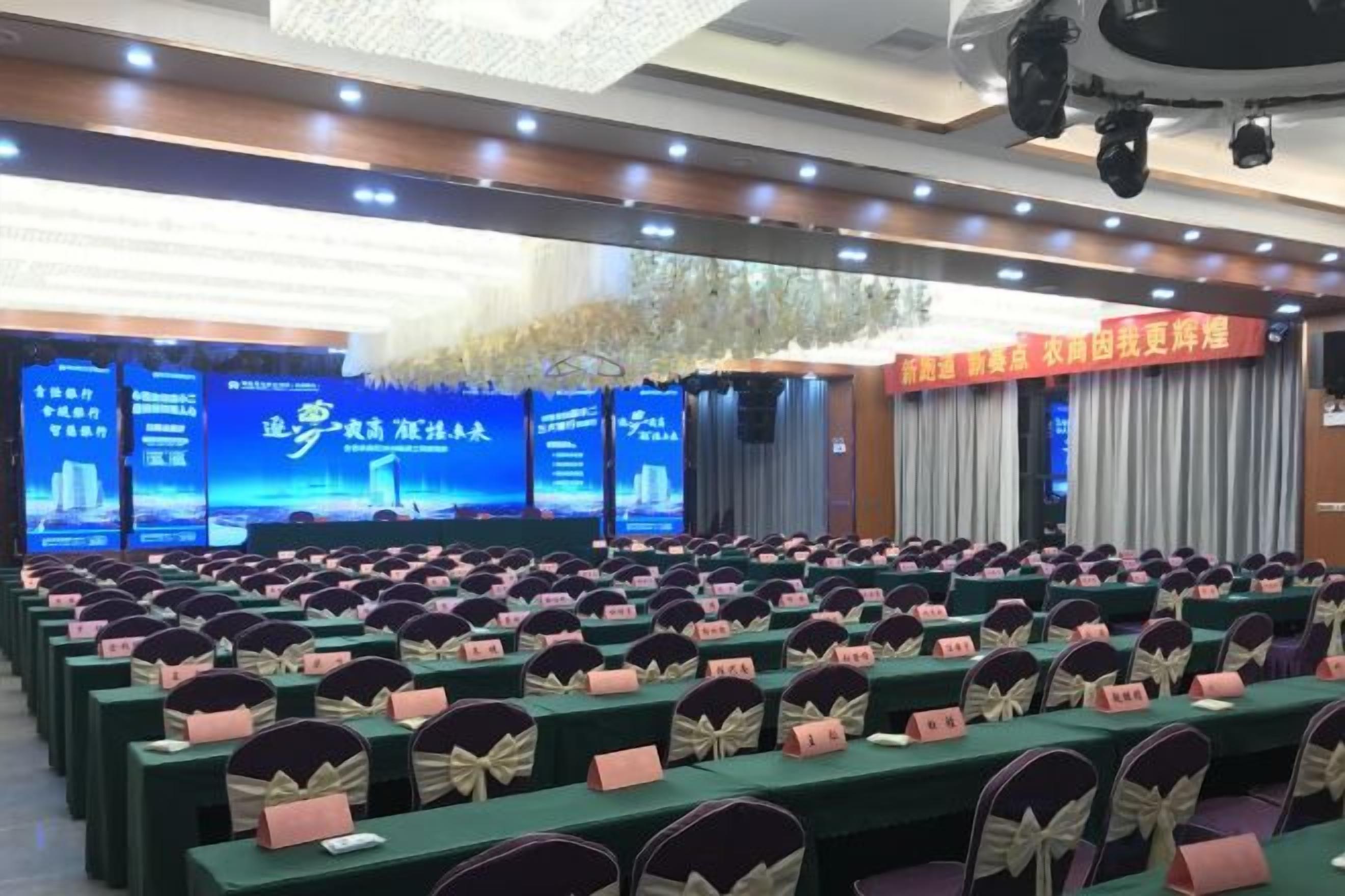 Un salón de conferencias con filas de mesas y sillas, un escenario con un telón de fondo azul y un cartel con texto.