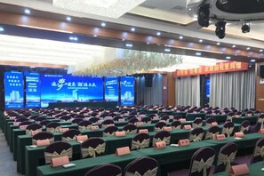 Un salón de conferencias con filas de mesas y sillas, un escenario con un telón de fondo azul y un cartel con texto.