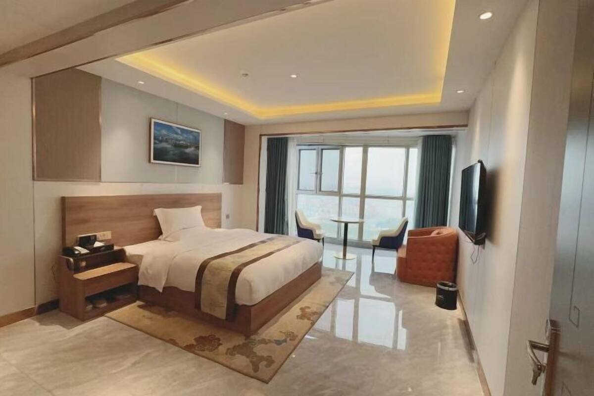 A modern hotel room with a large bed, a desk, a chair, a TV, and a balcony with a view.