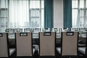 Un salón de conferencias con filas de mesas y sillas, ventanas amplias con cortinas y vistas a la ciudad.