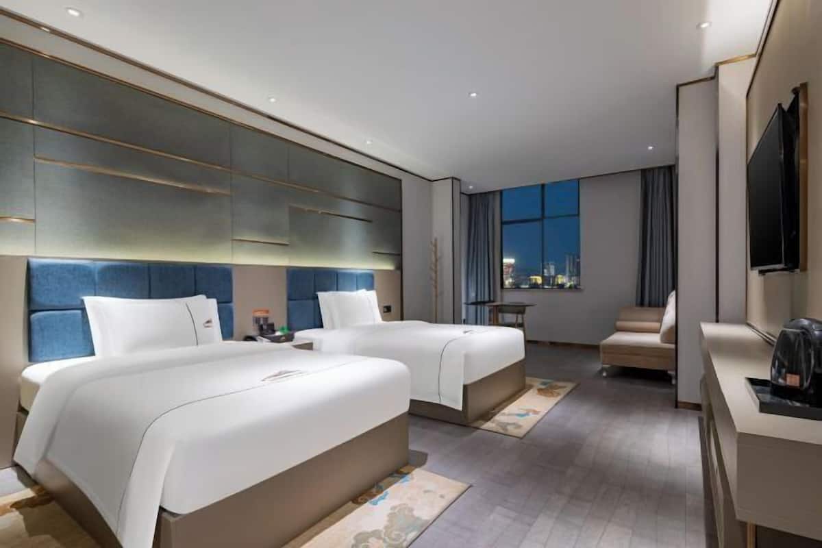 A hotel room with two beds, a desk, a chair, a TV, and a window with a city view.