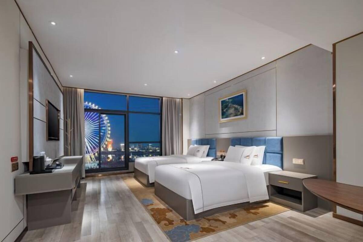 A modern hotel room with a large bed, a desk, and a view of a Ferris wheel at night.