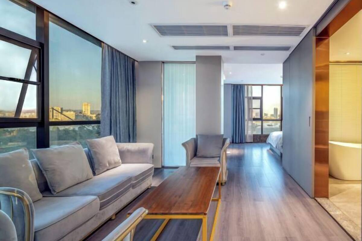 A modern hotel room with a sofa, armchairs, a wooden coffee table, and large windows offering a city view.