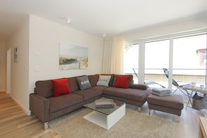 A modern living room with a brown sectional sofa, a glass coffee table, and a view of the outdoors through large windows. - Villa Stern Apartment 5 - Villa Stern (Timmendorfer Strand)