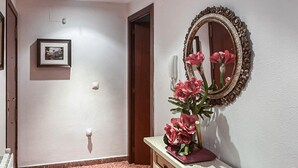 Un couloir avec un tableau encadré au mur, un miroir avec un cadre floral et un vase rempli de fleurs posé sur un comptoir.