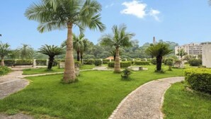 Un jardín arbolado con palmeras, un sendero de grava y un edificio al fondo.