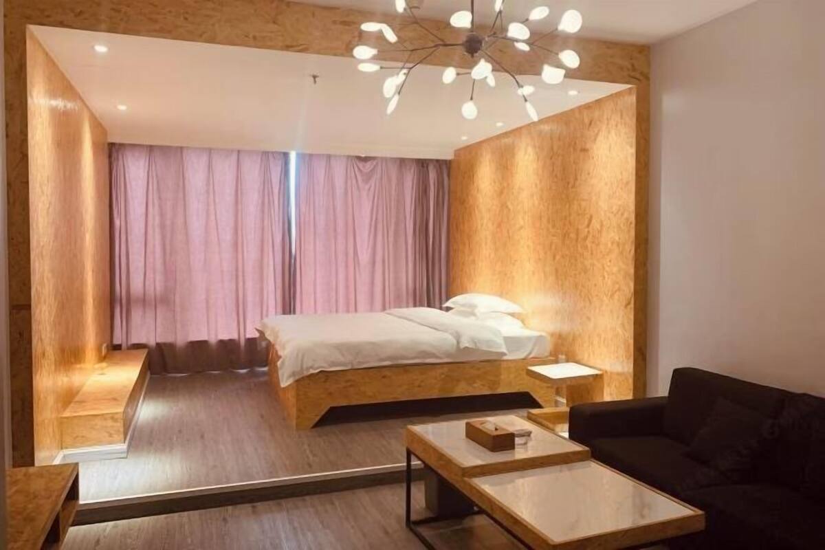 Habitació d'hotel moderna amb llit de fusta, un sofà negre, taula de centre de fusta i una finestra gran amb cortines.