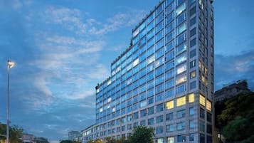 Un hotel de varios pisos con una fachada moderna de vidrio, situado en un entorno urbano con farolas visibles y otros edificios en el fondo.